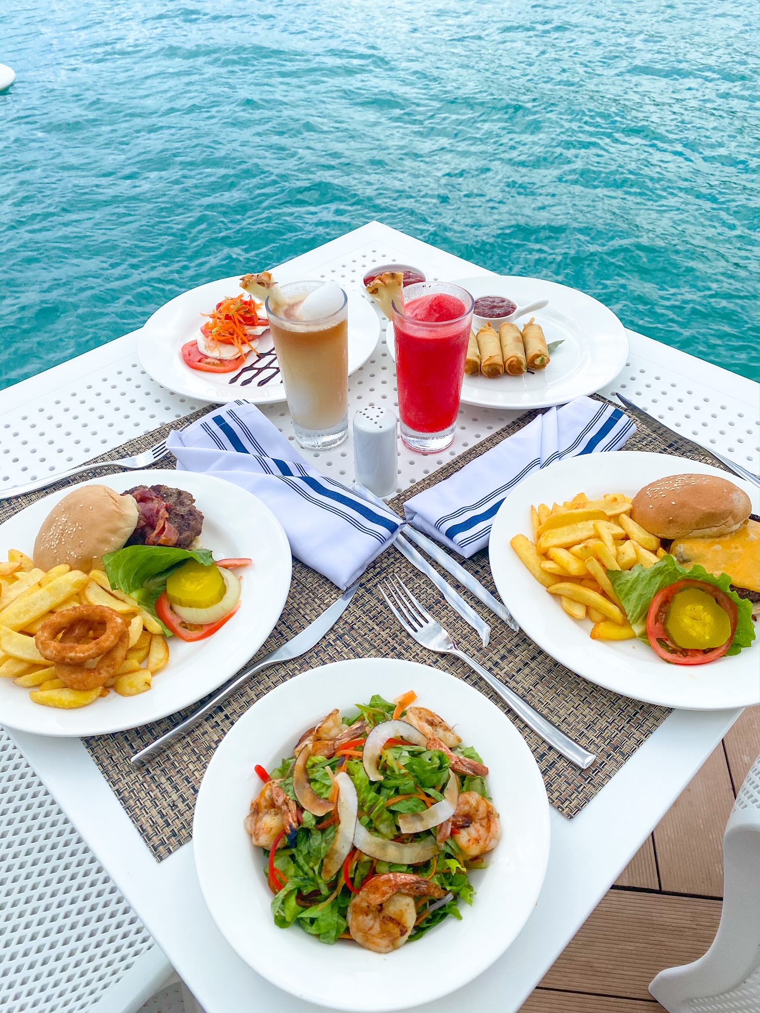 Several plates of food and a few drinks on a white table with the ocean in the background