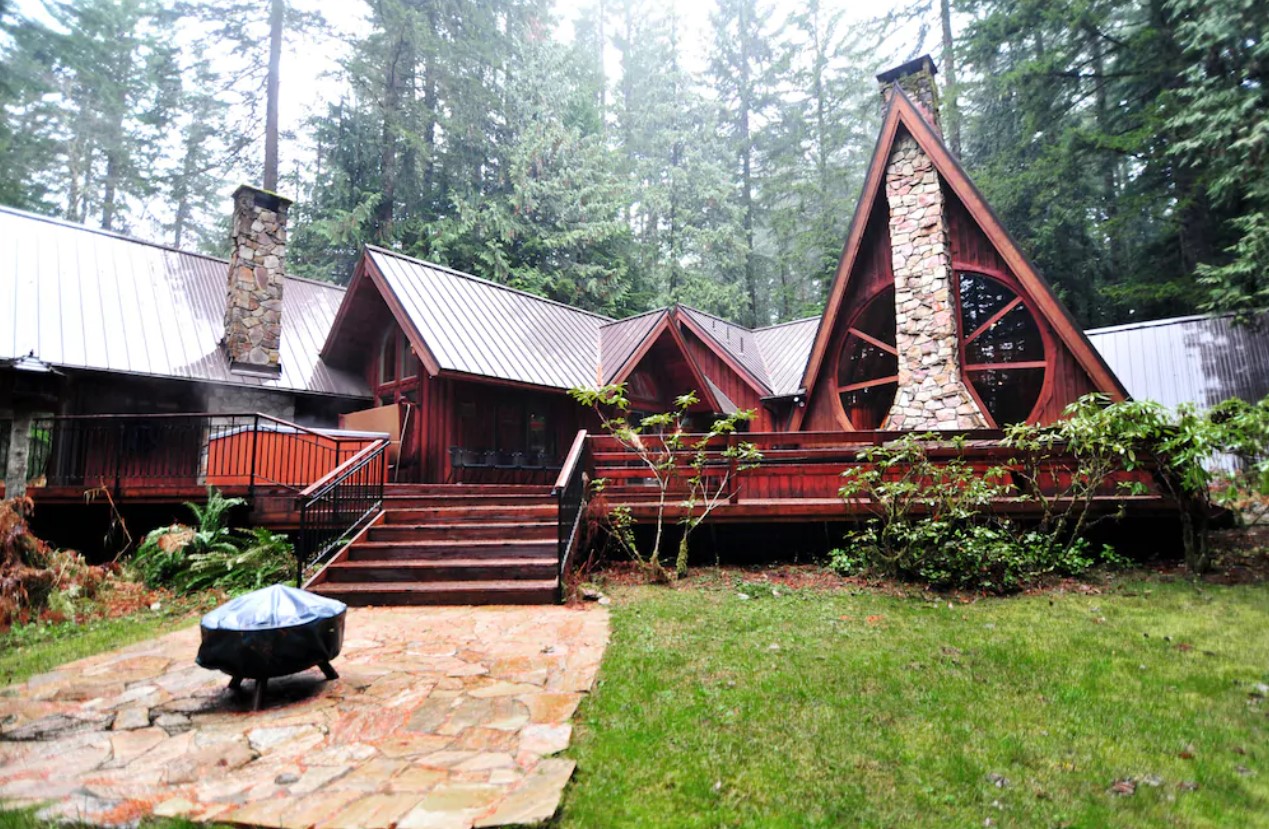 A large dark red lodge in the woods with several peaks, and two large semi-circle windows around a stone chimney. There is also a large deck, an outdoor fire pit, and plenty of trees