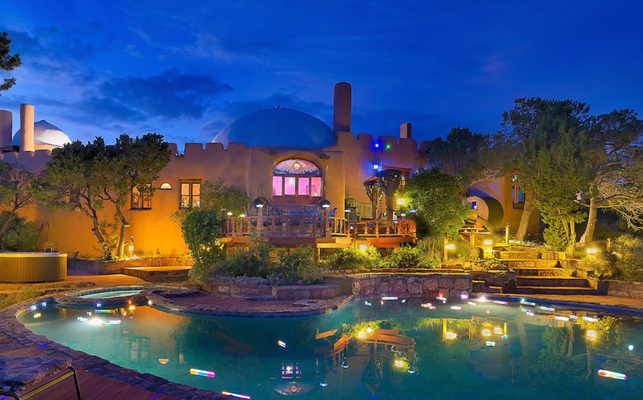 A large home with a domed ceiling, stained glass windows, a massive pool with lights in it, a green space with stone steps and trees, and more at twilight.