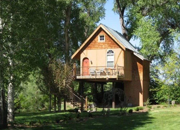 A cute fairy tale cottage style treehouse with cedar shingling, a curved orange door, and steps that lead up to the treehouse and deck. It is surrounded by grass and tall trees