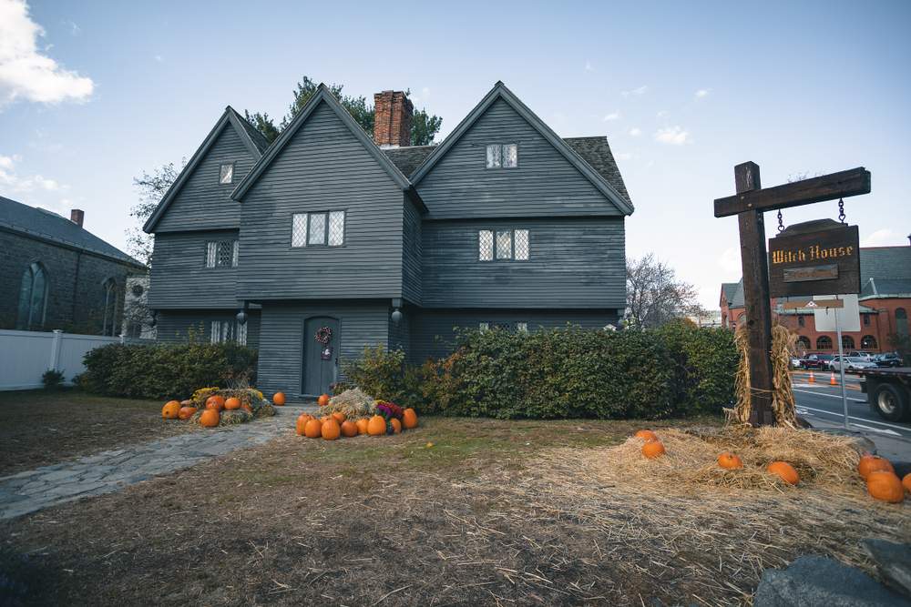The infamous Witch House in Salem Massachusettes. It is a pre-colonial puritan home that is painted all black and has three peaks. There are small windows on it. In front of it are piles of pumpkins and straw.