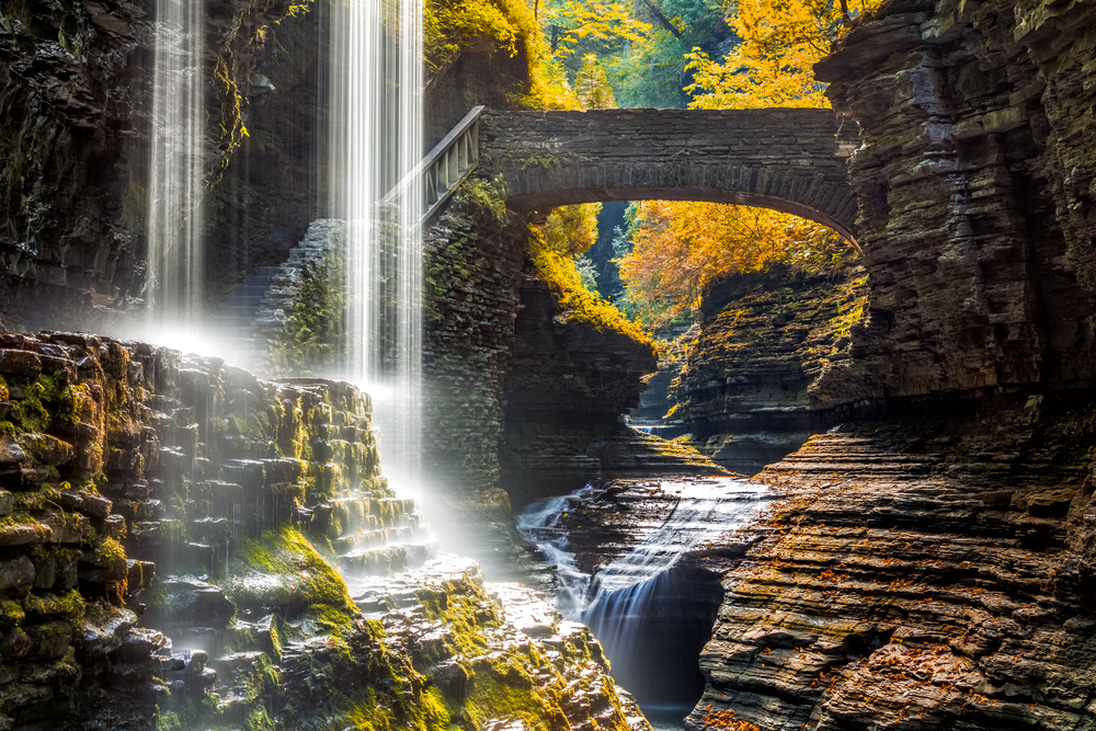 A waterfall falling down into a ravine with a stone bridge and staircase. There are trees with yellow leaves behind the rock formations and moss growing on the rocks.