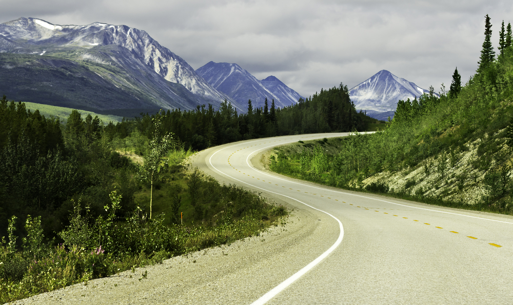 A winding road in Alaska with tall snow topped mountains in the distance. The road is surrounded by greenery and large trees. It is cloudy. 