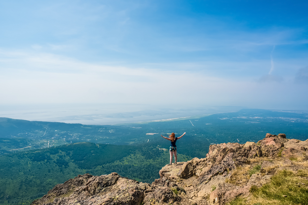 A woman with her hands spread wide on the edge of rocky cliff looking out to a valley. The valley is very green and has rolling hills and is in Anchorage Alaska. It is one of the best bachelorette party destinations.