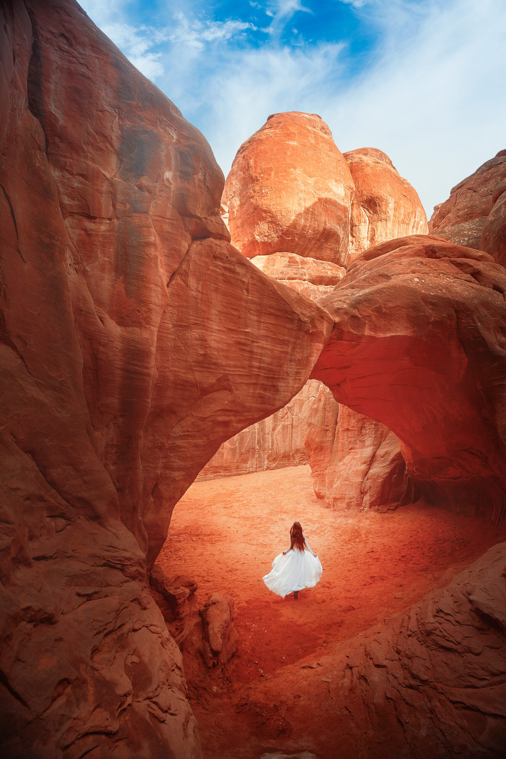 A woman in a white dress with long hair running under a red sandstone arch. The area is nothing but red sandstone rock formations. Its a great Southwest road trip stop.