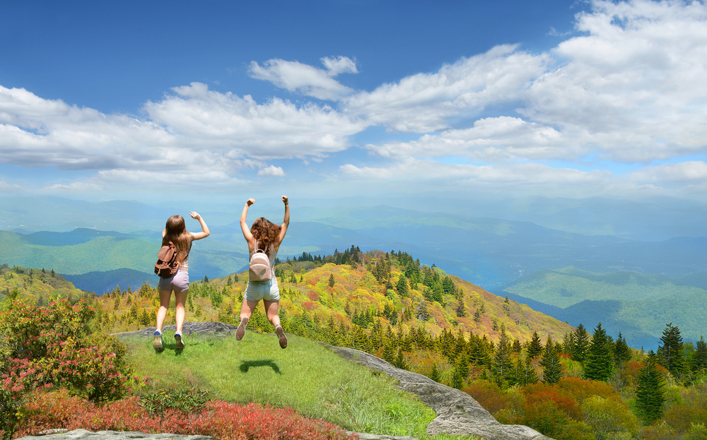 Two women jumping on the side of a mountain in Asheville North Carolina. They are facing the rolling mountains where you can see trees, valleys, and clouds shadowing the ground.