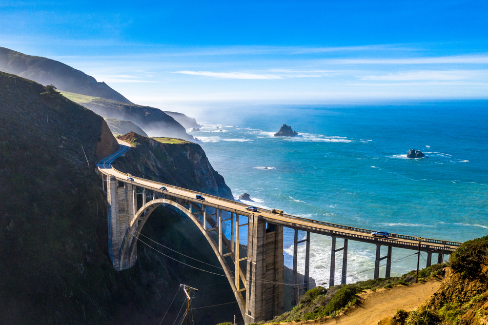 A side aerial view of the Bixby Bridge on a sunny day. You can see a few cars on the bridge and the pacific ocean with rock formations behind it. The mountains that the bridge leads to are in shadow. It is one of the most popular stops on any Big Sur road trip