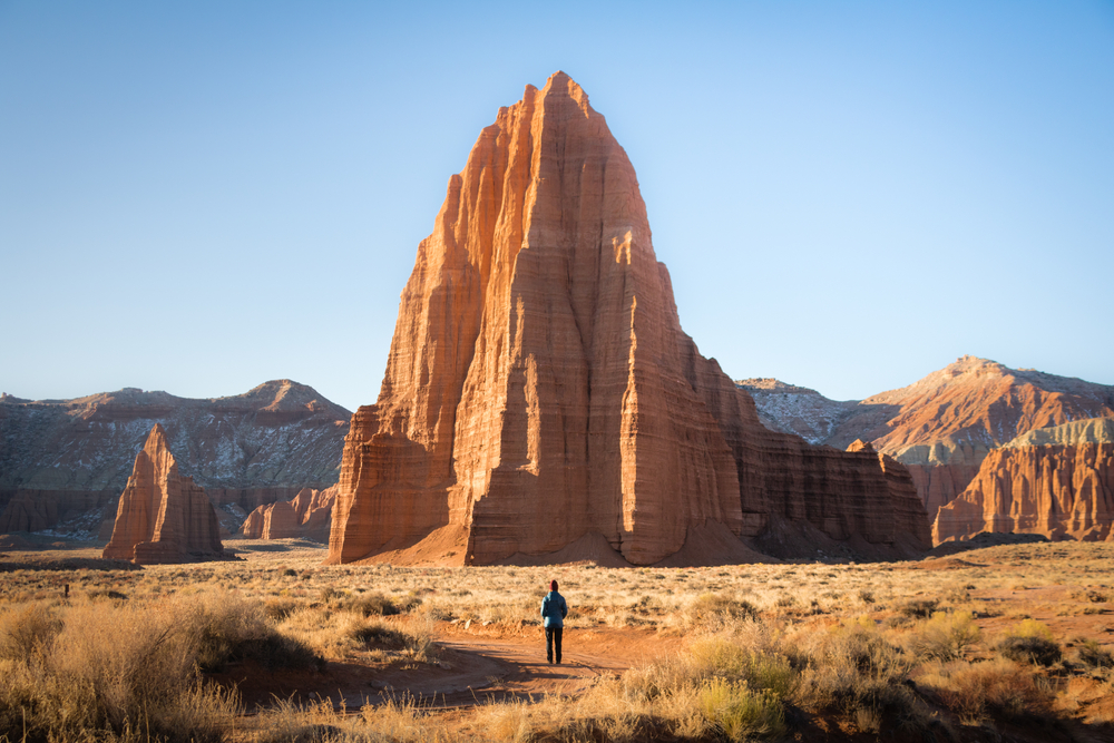 The large Temple Sun rock formation at Capitol Reef National Park. It is a light orange rock formation that is taller than the other mountains and rocks near it. There is a person in dark clothes standing in front of it and they look very small.