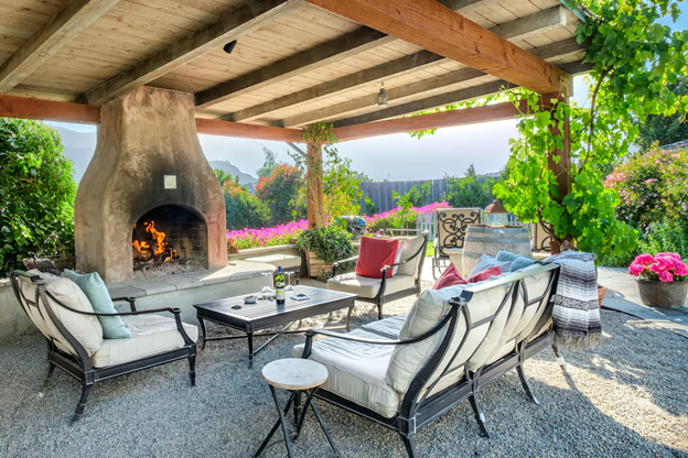 The private patio with seating, a fireplace, and covered pergola with vines growing on it. There are pots of pink flowers around the patio.