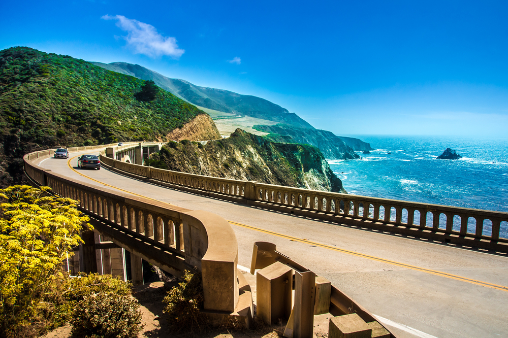 A portion of Big Sur that goes over the water and winds towards a rocky mountain. You can see the pacific ocean with rock formations on one side and mountains on the other. 