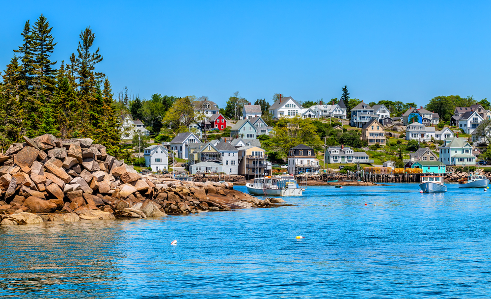 Looking at a charming town from the water in Coastal Maine. The town is right on the water and has colorful homes and boats in the water. You can also see some of the rocky shore with trees on it. 