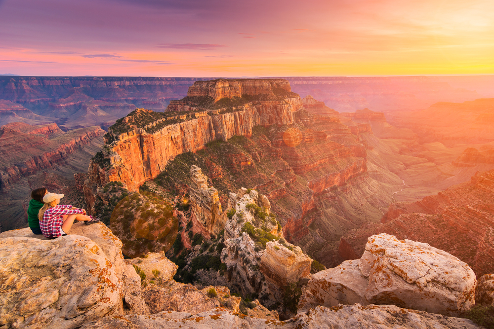 A couple sitting on a rock looking out at the north ridge of the Grand Canyon. It is sunset and the sky is orange, yellow, pink, and purple. A great Southwest road trip stop.