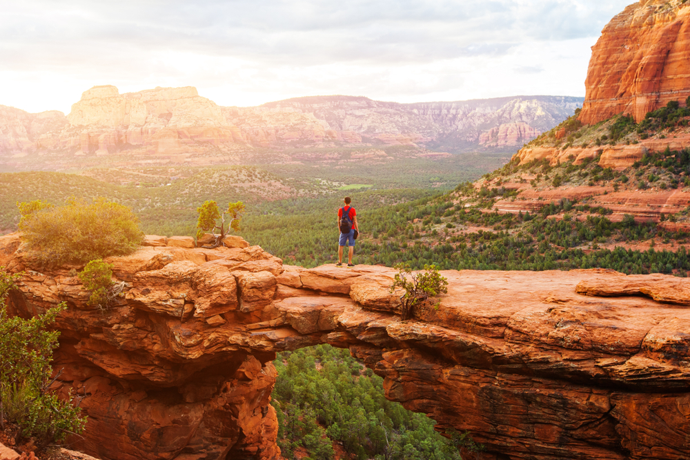 A person in a red shirt and blue shorts standing on the Devil's Bridge trail in Sedona Arizona. It is a red sandstone bridge rock formation that looks out onto a valley with trees and sandstone mountains in the distance. 