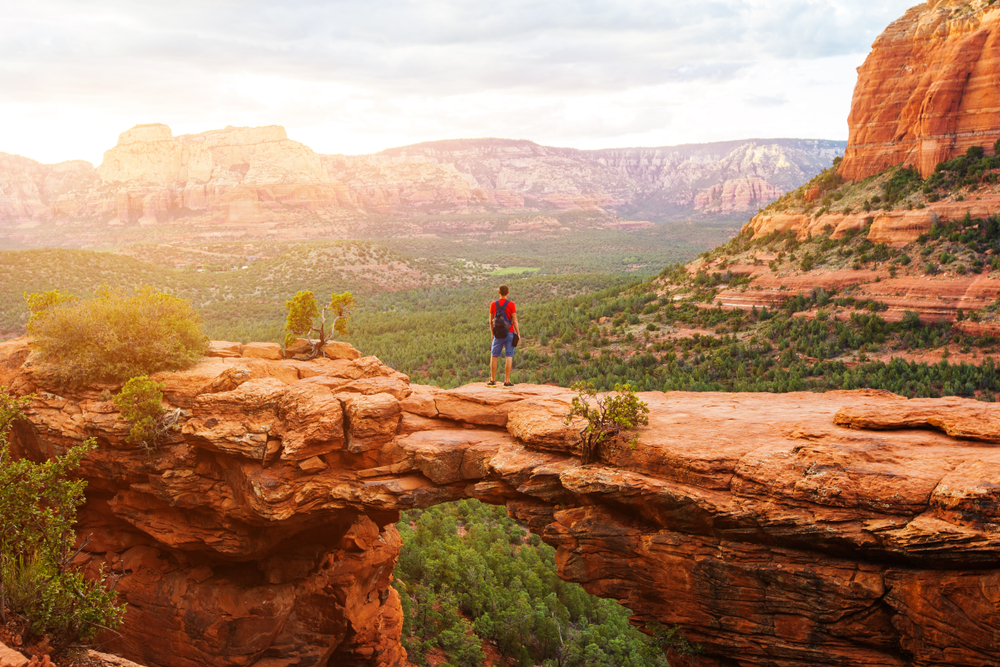 A person in a red shirt and blue shorts standing on the Devil's Bridge trail in Sedona Arizona. It is a red sandstone bridge rock formation that looks out onto a valley with trees and sandstone mountains in the distance.