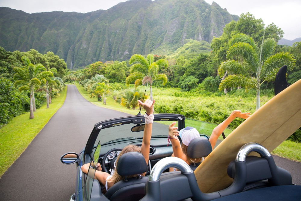 Two women with their hands up riding in a car with the top down and a surf board in the back seat. They are driving on a road in Hawaii surrounded by greenery and palm trees. In the distance you can see a large tropical style mountains. One of the best bachelorette party destinations.