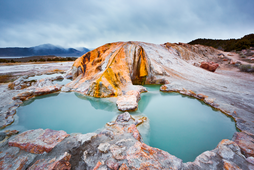 A natural hot spring in a rock formation. The area is full of smooth rocks and you can see mountains and trees in the distance. The water in the hot spring is a teal blue and there is steam in the air. 