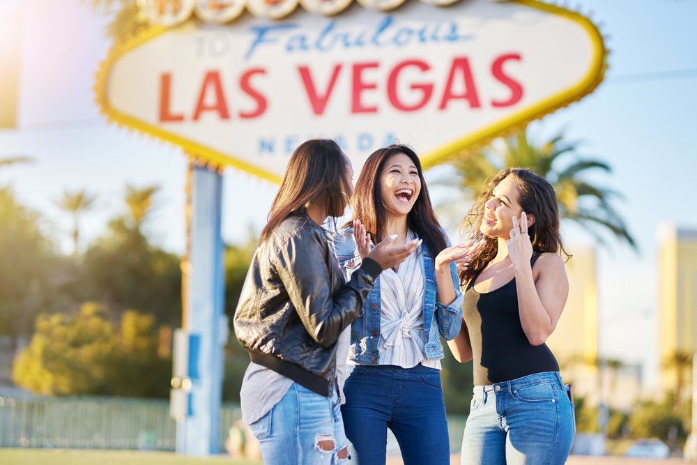 A group of three women laughing in front of the famous Las Vegas sign in Las Vegas. They are smiling and laughing with each other. 