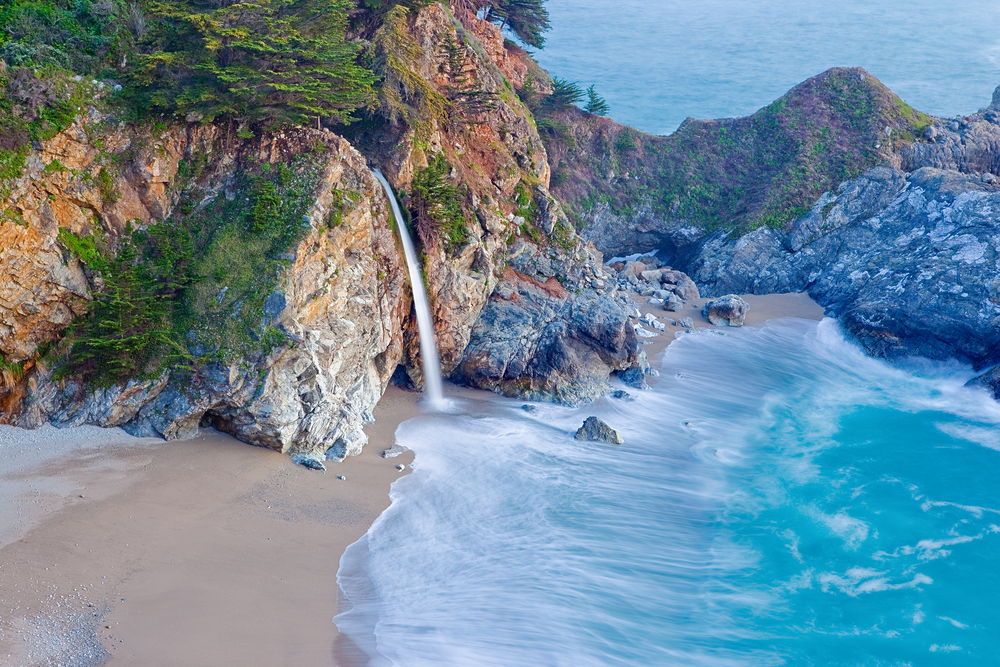 An aerial shot of the popular McWay falls, a large waterfall that flows directly onto a beach. The waterfalls comes from a large rock formation and the sand is pale tan and the water is a beautiful crystal blue. 