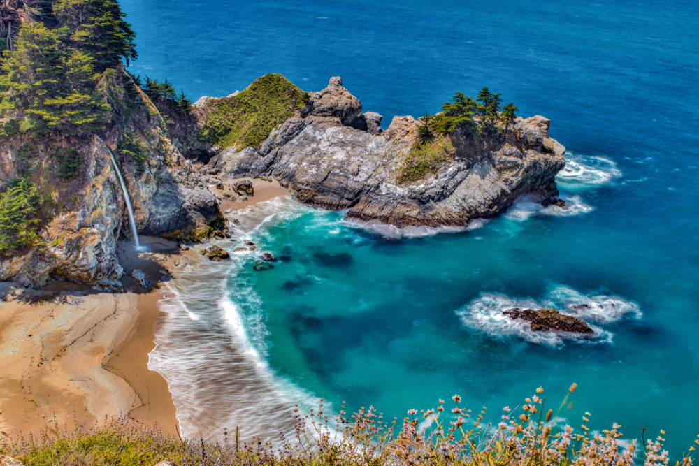 The view over the rocky cliffs to McWay Falls Cove. You can see the waterfall, large rock formations with trees growing on them, and the crystal blue pacific ocean.