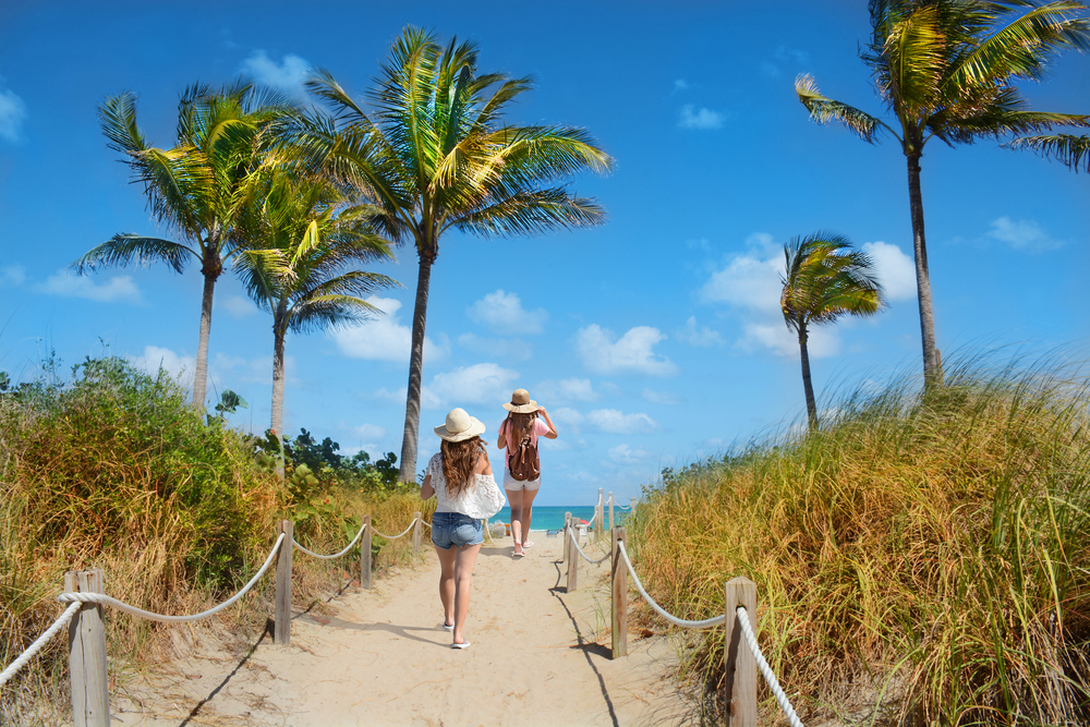 Two women in shorts and sun hats walking on a sandy path over the sand dunes towards the beach in Miami. The beach has large palm trees and you can see a glimpse of the crystal blue water