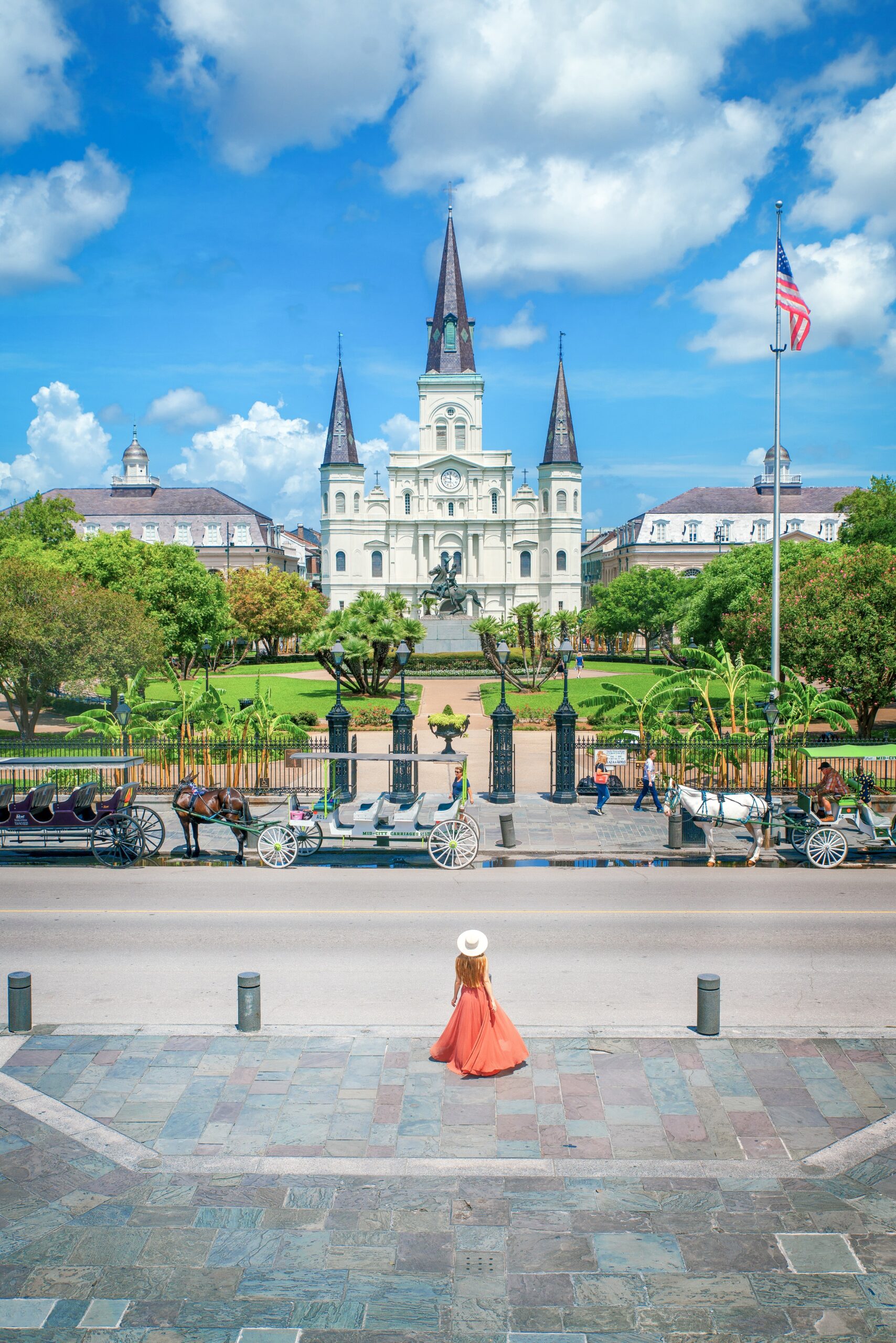 A woman in an orange maxi dress and white sunhat standing in Jackson Square in New Orleans on a sunny day.