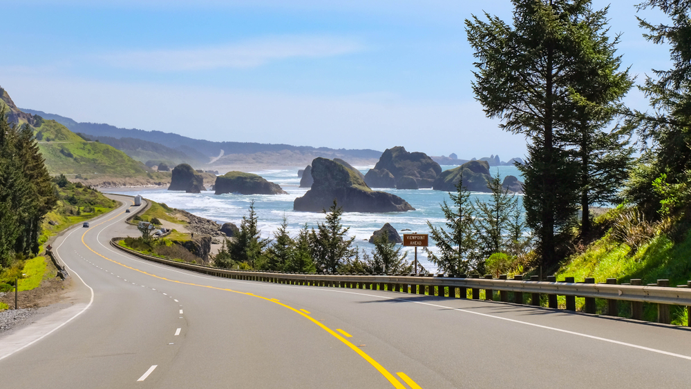 A highway on the Oregon coast. You can see large rock formations in the ocean and on the other side of the highway are green mountains. One of the best west coast road trip ideas. 