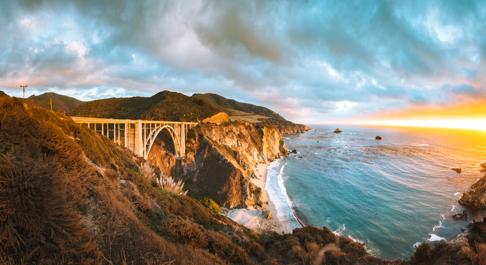 A panoramic of the Bixby Bridge at sunset. You can see the bridge, the rocky cliff side, and the pacific ocean. The sky is cloudy and the sun is bright orange. A popular stop on a Big Sur road trip