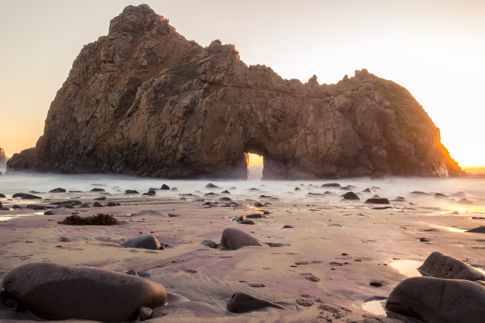 A large rock formation with a large door keyhole almost right in the middle of it. Around the rock formation is sand with large stones and puddles of water in it. There is a light fog on the ground and it is sunrise, so the sun is coming up behind the rock formation.