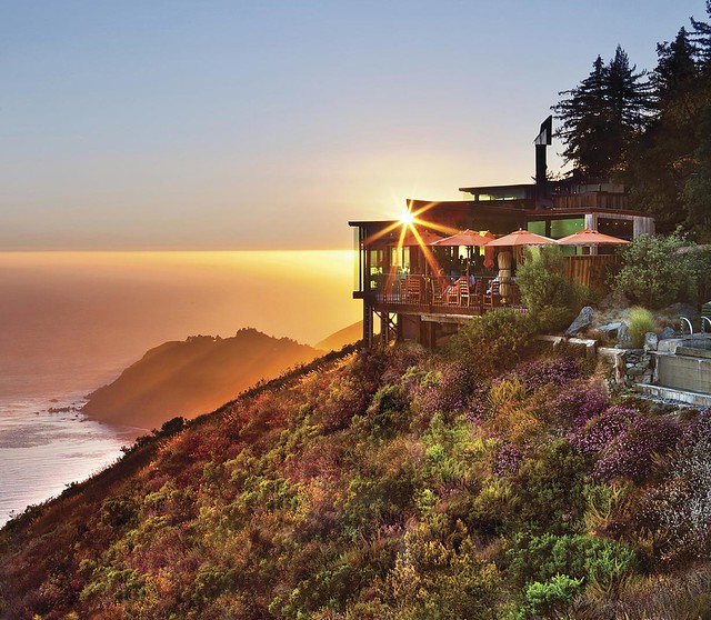 A luxury building on the side of grassy cliff over the Pacific Ocean. It is sunset, and behind the cliff you can see other rock formations. The building has a deck with patio tables and umbrellas on it and lots of windows. 