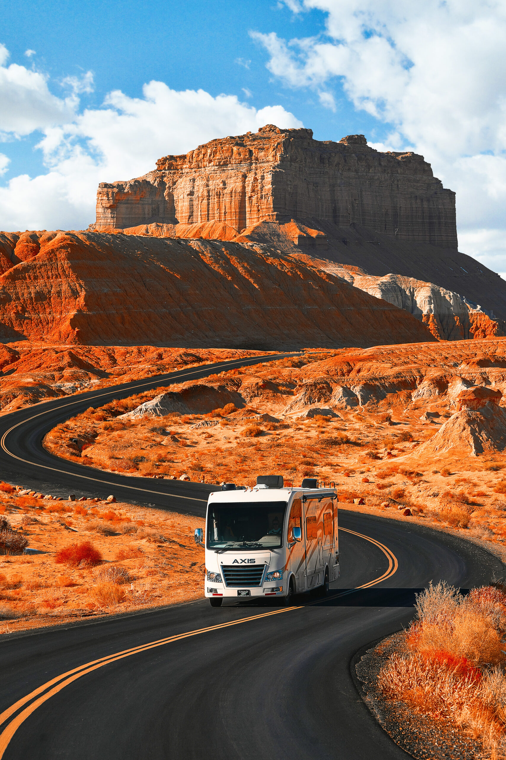 An RV driving through the red sandstone of the Southwest. In the background you can see a hill with a large red sandstone rock formation on top of it. 