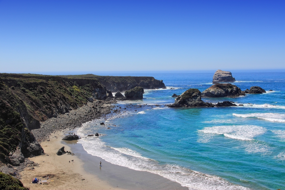 The view of the shore of Sand Dollar Beach in Big Sur from the top of the rocky cliffs. The shore is fairly sandy but there are large rocks on it. The ocean is bright blue and there are large rock formations in the water. 