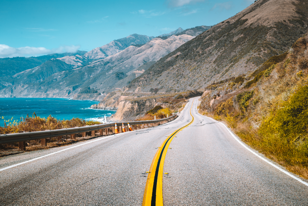 A scenic road on the coast of California. On one side of the road you can see a rocky shoreline and the pacific ocean. On the other side of the road are mountains that look very dry, almost like desert mountains. 