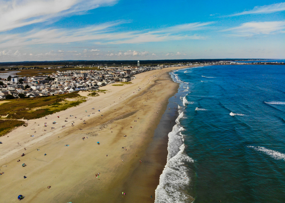 An aerial view of the shore of the Hamptons in Long Island. You can see people relaxing on the beach, large waves in the ocean, and homes behind the sandy shore. One of the best bachelorette party destinations.