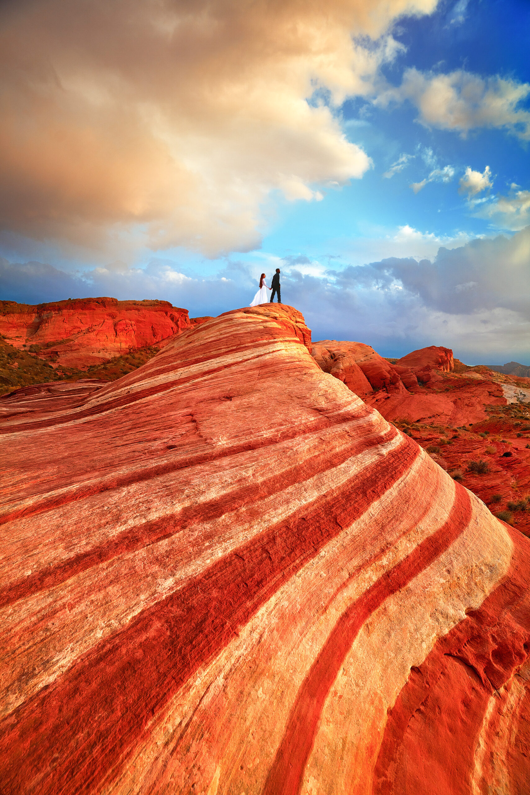 A woman in a white dress holding hands with a man in all black standing on a red and orange striped rock formation. The sky is blue and there are big fluffy clouds. 