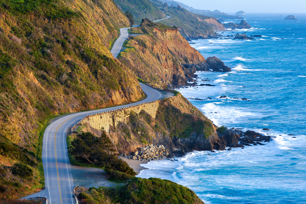 A view of Big Sur winding on the sides of the cliffs in the distance. On one side of the road is the pacific ocean and on the other side are rocky mountains with greenery. Part of the Big Sur road trip