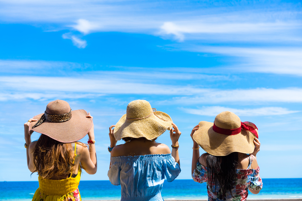 Three women standing next to each other wearing sundresses and sun hats. They have their hands on their sunhats and are looking out onto a bright blue ocean and a bright blue sky.
