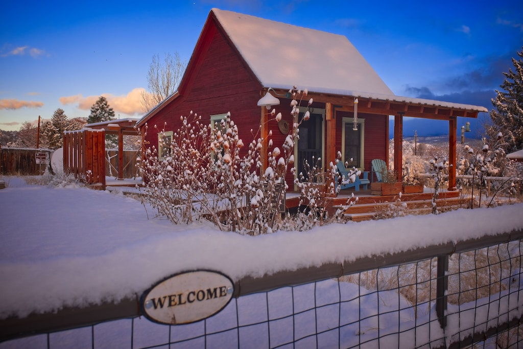 A view of the charming red pioneer cabin, one of the best VRBOs in Utah. Snow has fallen and it is sunset, adding to the quaint farm charm of the Pioneer Cabin.