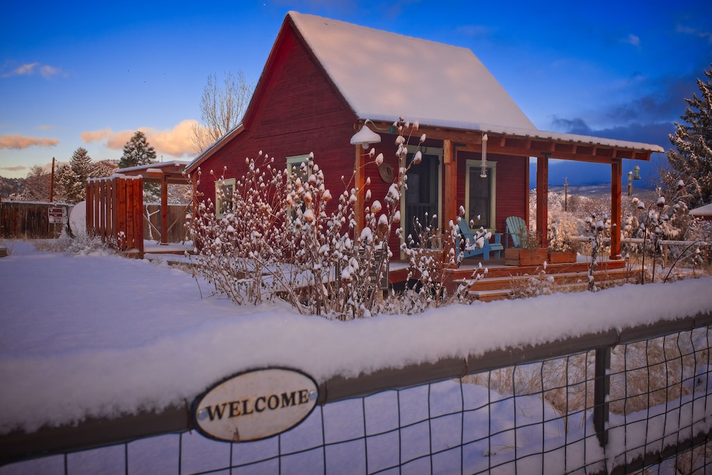 A view of the charming red pioneer cabin, one of the best VRBOs in Utah. Snow has fallen and it is sunset, adding to the quaint farm charm of the Pioneer Cabin. 