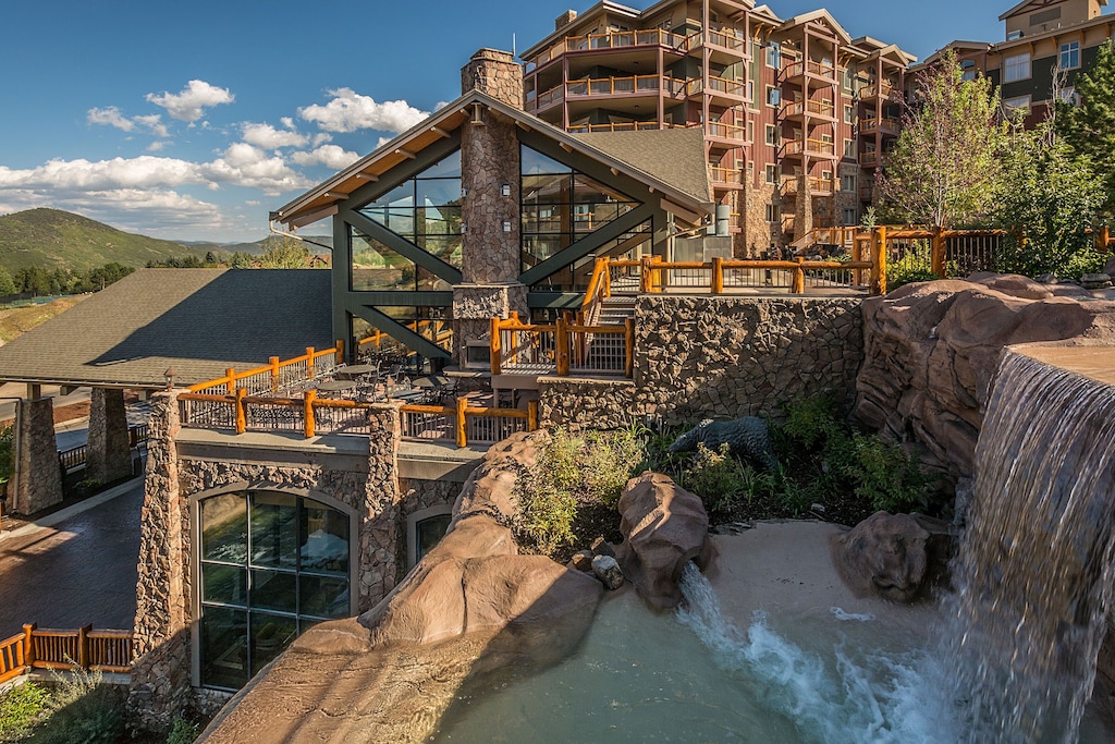 External view of the Westgate Park City resort one of the best vacation rentals in Utah. The view includes the mountains in the background and a manmade large waterfall in the foreground. 