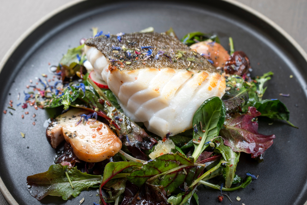 Cooked cod with spices and dried flowers sprinkled on it. It is sitting on a bed of greens with a type of dressing drizzled on it. Everything is on a black plate. 