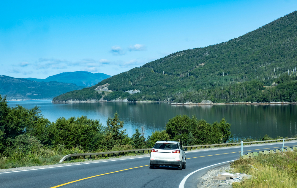 A white car driving on a road that is on the shore of a river or bay. There are hills on the shore of the water that are covered in trees. The sky is clear and blue. 