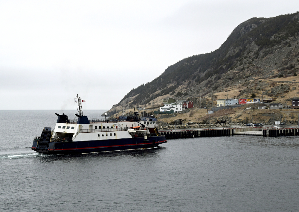 A black and white ferry pulling into a dock on the shore Newfoundland. There is a hill with colorful houses on it. It looks like it may be winter as nothing is growing and it looks kind of dreary.