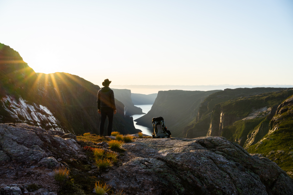 A person standing on a cliff wearing a hat looking out onto a mountain landscape. There is also a backpack next to them on the cliff. The sun is beginning to set behind one of the mountains. 