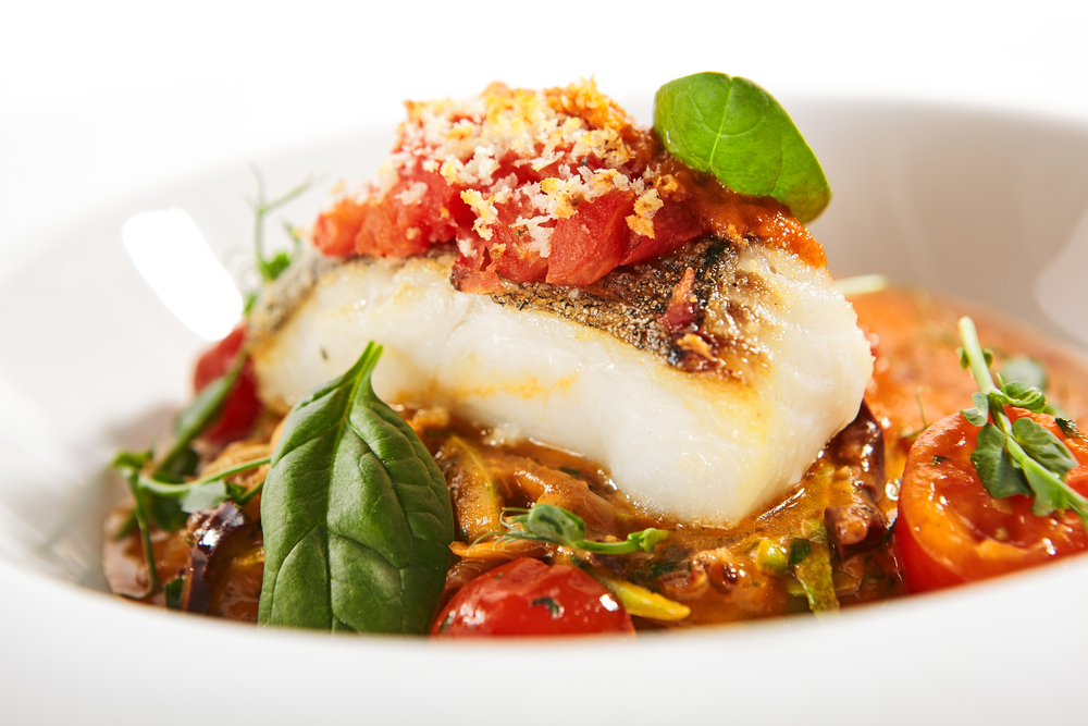 A white plate of cooked halibut. It is sitting in a creamy red sauce with sliced tomatoes, basil, and breadcrumbs sprinkled on top.