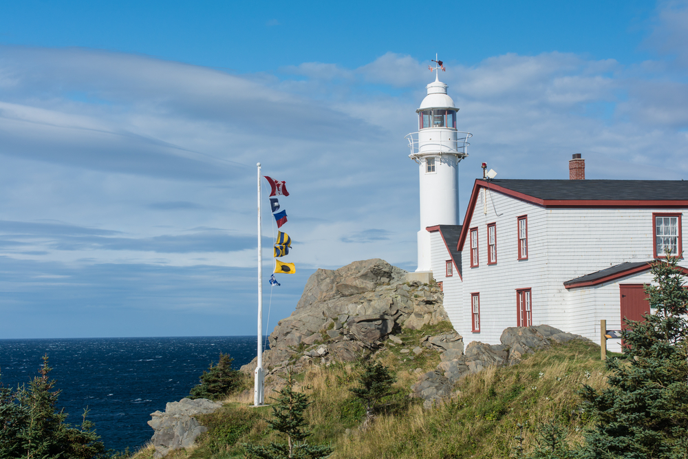 A white lighthouse with red trim looking over the shore at Lobster Cove. There are large rock formations, grasses growing on the rocks, and a flag pole with different types of flags on it. You can just see the water over the rocks. 