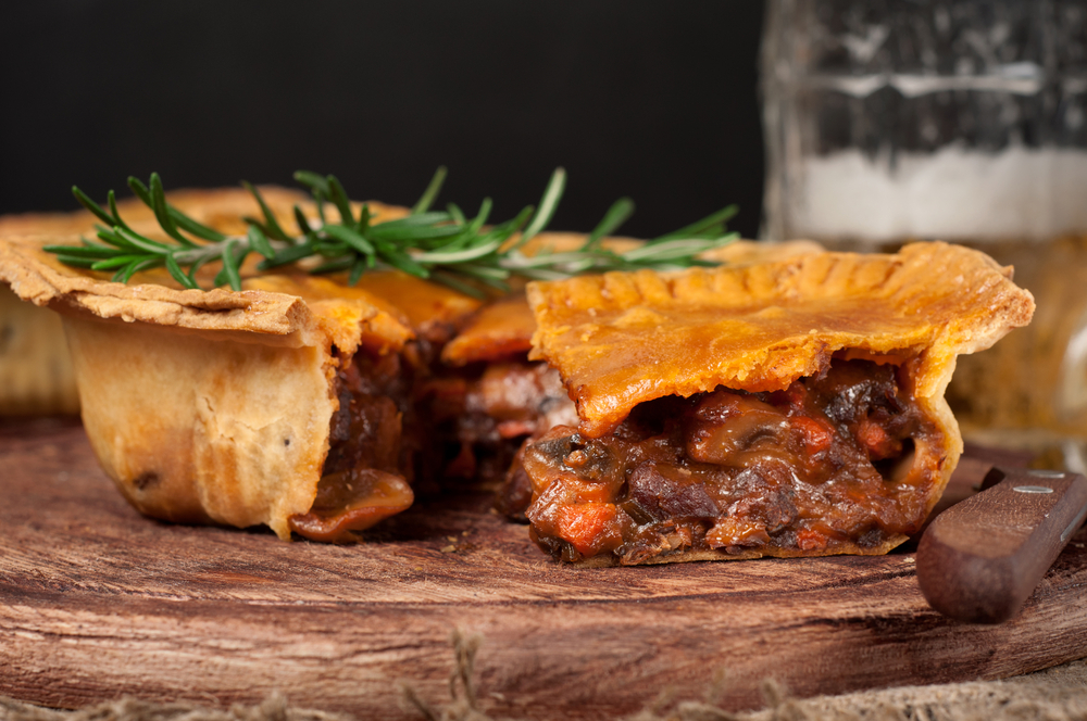 A meat pie that is cut open on a wooden board. You can see beef, carrots, mushrooms, and sauce in the pie. On top of it is a sprig of rosemary. 