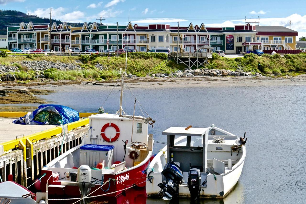 Looking at a large multicolored hotel on a bay in West Newfoundland. It is right on the shore and you can see tall grassy dunes and rocks in the shore. There are also a few boats tied up to a dock across from the hotel. 