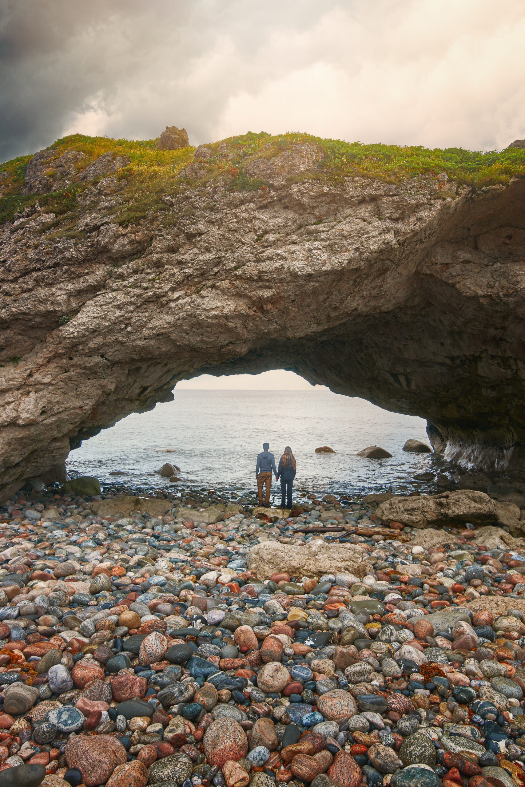 A couple standing holding hands under a large natural stone archway. They are looking out to the water. The shore of the area is nothing but rocks in all shapes, sizes and colors. 