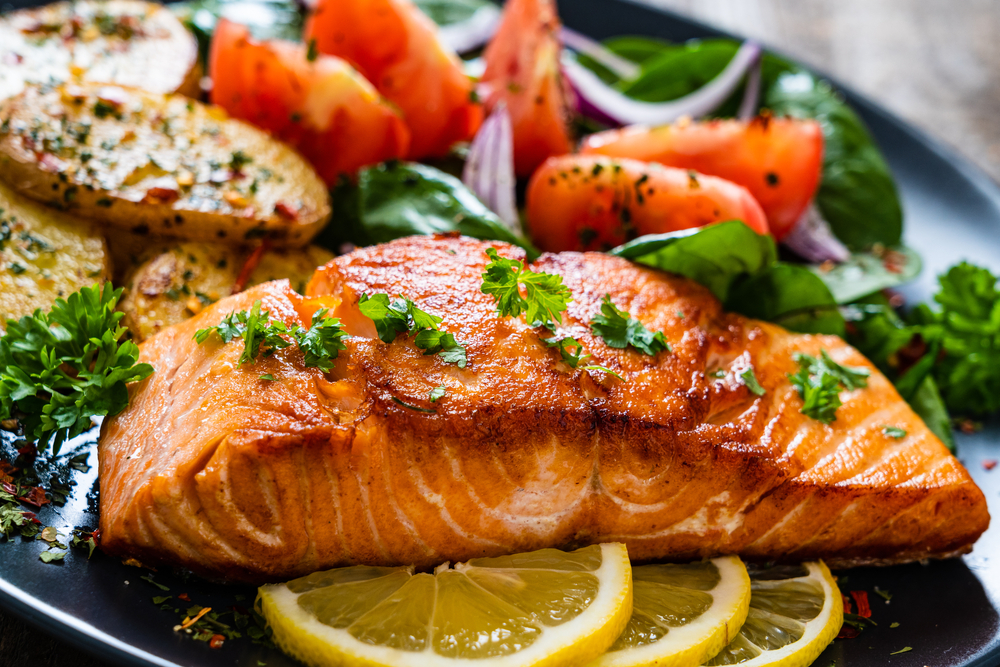 A plate of a salmon dinner. The salmon looks grilled and is sitting on sliced lemon. You can also see a salad and wedge baked potatoes on the plate. 