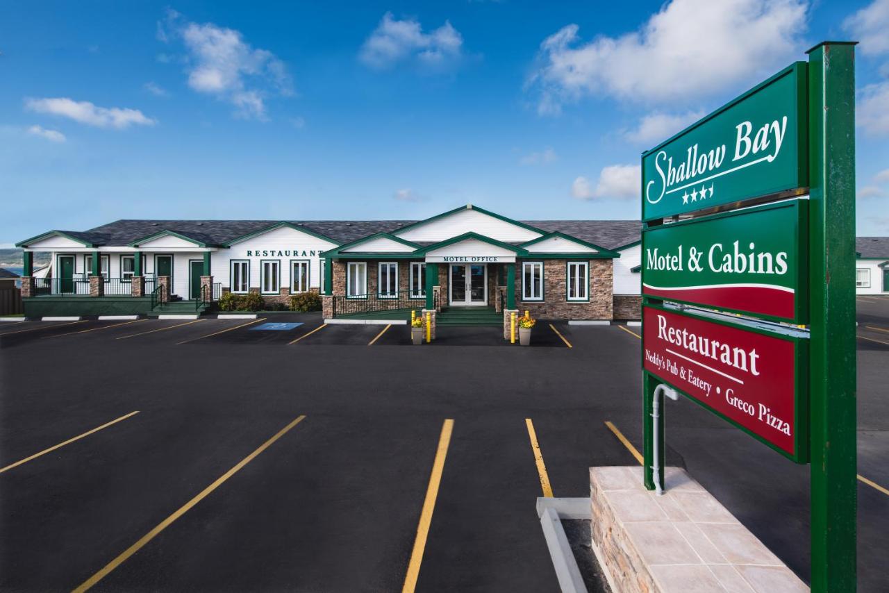 The front of the Shallow Bay Motel. It is a stone and wood motel. The wood is painted white with green trim. In front of it is a large parking lot. There is also a sign that has the motel name and advertising a restaurant. The sign is green and red. 